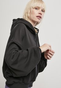 Felpa nera oversize con un ampio cappuccio, maniche voluminose, orlo elasticizzato e texture morbida. Nessun motivo visibile o dettagli in metallo.