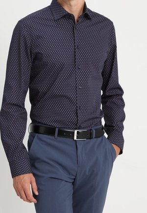 Homme portant une chemise à manches longues bleu foncé à motif, rentrée dans un pantalon bleu clair avec une ceinture noire, main dans la poche.