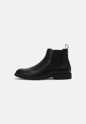 Botte Chelsea en cuir noir avec panneau latéral élastique et languette sur fond gris clair uni.
