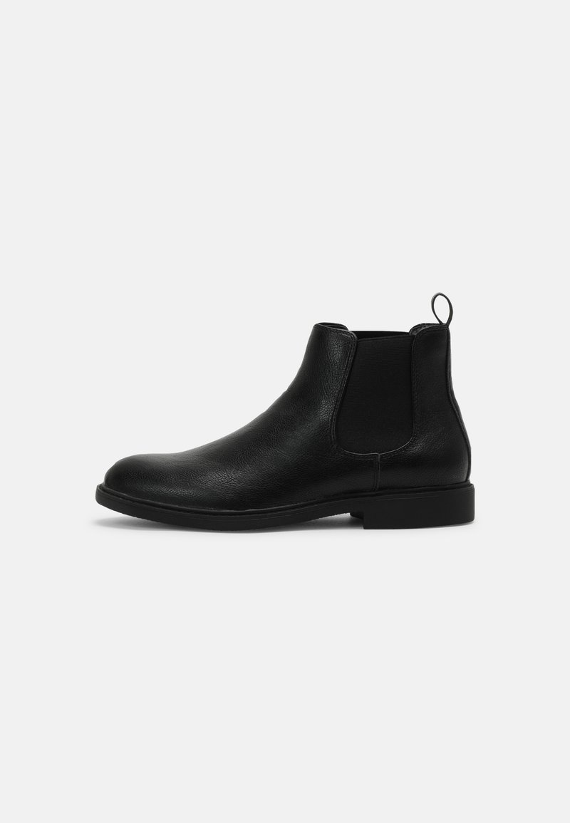 Botte Chelsea en cuir noir avec panneau latéral élastique et languette sur fond gris clair uni.