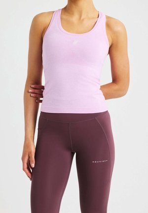 Kvinde iført ærmeløs lyserød sportstop og højtaljede mørke mauve leggings med sidelomme, stående med den ene arm bag ryggen.