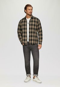 Geruite flanellen overhem met zwart en beige, met knopen aan de voorkant, een wit t-shirt eronder en een donkere spijkerbroek. Draagt witte sneakers.