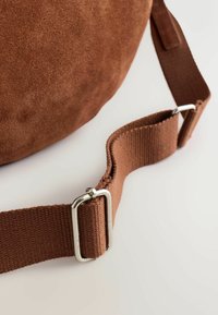 Braune Crossbody-Tasche aus Wildleder mit strukturierter Oberfläche und einem gurtartigen Stoffriemen mit Metallschließe. Runde Form mit minimalen Details.