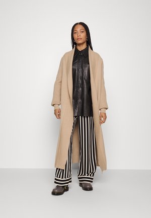Beige gestrickte lange Strickjacke, schwarzes Lederhemd mit Knöpfen, schwarz-weiß gestreifte weit geschnittene Hose und graue Clogs mit Schnallen.