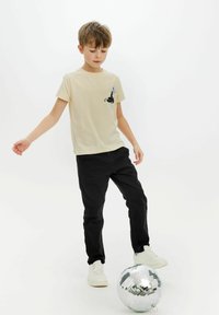 Camiseta beige con un diseño gráfico negro en el pecho, combinada con pantalones negros y zapatillas blancas. Bola de discoteca reflectante cercana.