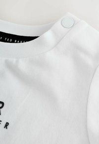 Valge puuvillane T-särk ribakraega, millel on must trükitud logo ja dekoratiivne nööbikas detail õlal.