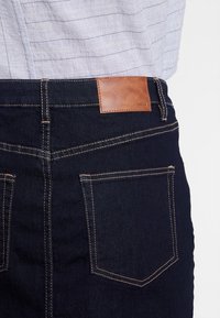 Tmavě modré džíny z denimu s hladkým povrchem, které mají na pásku hnědou koženou nášivku a viditelné švy na zadních kapsách.