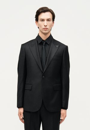 Jeune homme aux cheveux bruns courts, portant un costume noir ajusté, une chemise noire et une cravate noire, se tenant devant un fond clair uni.