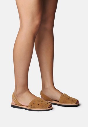 Nohy obuté v béžových sandálech na zadní patu s otevřenou špičkou, květinovými výřezy a černou podrážkou na bílém pozadí.