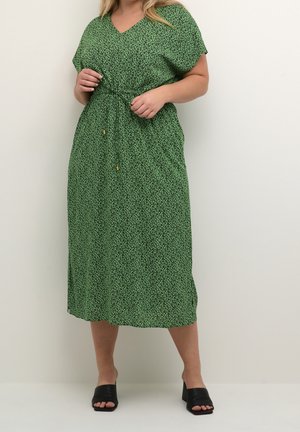 Donna che aggiusta il fiocco in vita su un vestito midi verde con piccolo motivo nero, abbinato a sandali neri con tacco largo e punta aperta su sfondo semplice.
