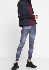 Tenue de sport comprenant une veste matelassée bleu marine avec des manches noires, un haut bordeaux et des leggings à motifs géométriques dans des tons de gris et de bleu.