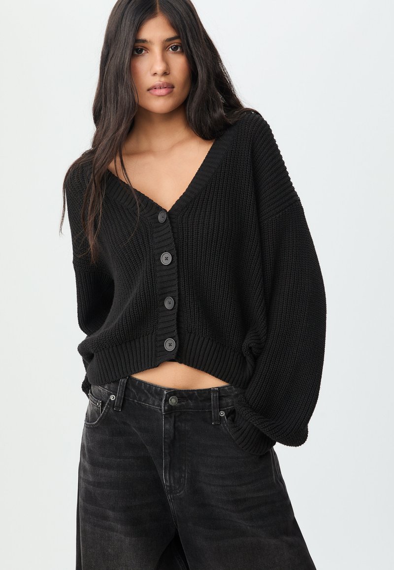 Schwarze, kurz geschnittene Strickjacke aus strukturiertem Strickstoff, mit tiefem V-Ausschnitt, sechs schwarzen Knöpfen und weiten Ärmeln. Über dunklen Jeans getragen.