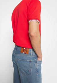 Camisa polo roja con detalles blancos, combinada con jeans de mezclilla azul claros que tienen un parche de cuero marrón marcado con "TH" y un detalle a rayas.