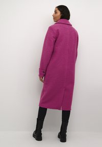 Lungo cappotto rosa con tessuto testurizzato, colletto a risvolto e polsini bottonati. Indossato sopra pantaloni neri e stivali neri fino al ginocchio.