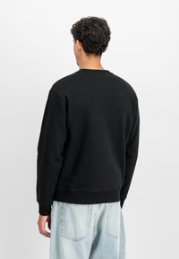 Melna kreklu ar apaļu kakla izgriezumu, kas izgatavots no kokvilnas maisījuma. Vienkāršs dizains, ribināts piedurkņu un apakšmalas. Modelis saskaņo ar gaiši ziliem platām biksēm, skatīts no aizmugures.