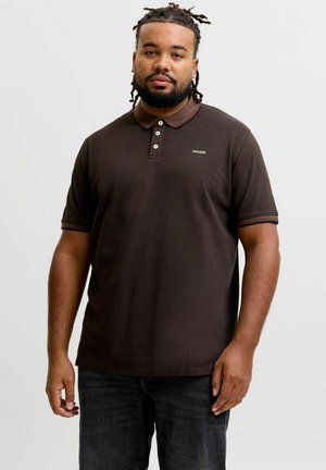 Homme aux cheveux tressés et à la barbe, portant un polo marron foncé et un jean noir, debout devant un fond clair uni.