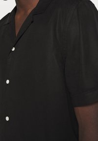 Chemise noire à manches courtes en tissu léger, avec un col classique et une fermeture à boutons sur le devant discrète. Texture lisse avec une coupe décontractée.