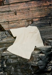 Kortärmad krämfärgad skjorta i texturerad tyg, med krage och knappstängning, mot en stenig bakgrund.