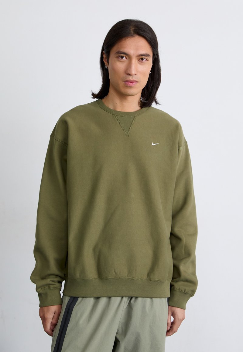 Oseba z dolgimi lasmi nosi oljčno zeleno jutranjo srajco z majhnim belim logotipom in stoji pred enostavno belo ozadjem.