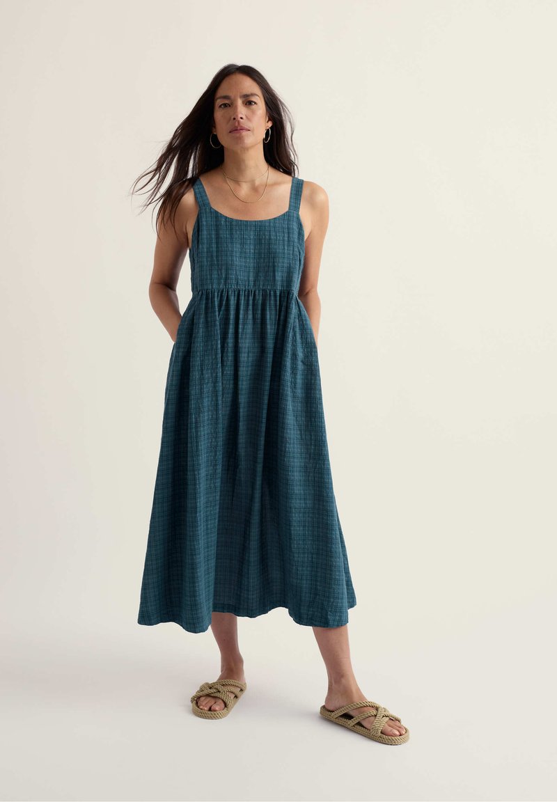 Femme debout, les mains dans les poches, portant une robe midi bleu sans manches et des sandales tressées beiges sur un fond neutre
