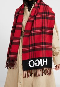 Femme portant un trench-coat beige et une grande écharpe à carreaux rouge et noir avec le logo "HUGO" et des extrémités à franges.