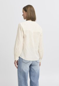 Blusa a righe color crema con maniche lunghe voluminose, orlo curvo e vestibilità rilassata, abbinata a jeans a vita alta di colore azzurro chiaro.