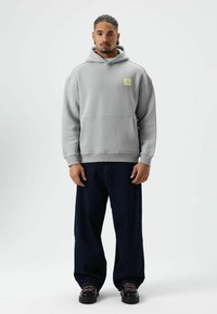 Grijze hoodie met een voorzak, geribbelde manchetten en een klein geel logo. Gecombineerd met losse donkerblauwe jeans en zwarte schoenen.