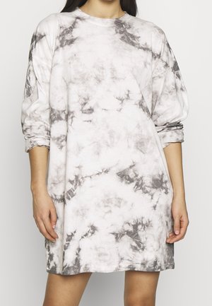 Femme portant une robe blanche à manches longues, longueur genou, avec un motif tie-dye gris, manches retroussées, debout devant un fond uni.