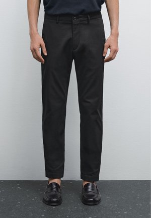 Black slim-fit trousers made from smooth cotton blend fabric. Features a belted waist, slanted pockets, and a cropped hem. Paired with black loafers.