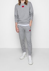 Sudadera y pantalones de chándal gris claro hechos de material suave, con parches de logo rojo. Los zapatos son blancos con un diseño texturizado.