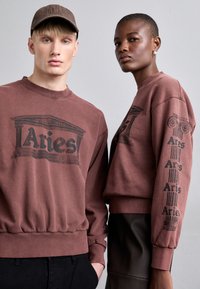 To personer bærer matchende bordeauxsweatshirts med et grafisk motiv af en klassisk søjle og teksten "Aries." Stoffet ser blødt og afslappet ud.