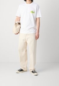 Valkoinen puuvillapaita, jossa on pieni värikäs graafinen kuvasto rinnassa, paritettu beigejen löysien housujen ja mustien matalavartisten tennareiden kanssa.