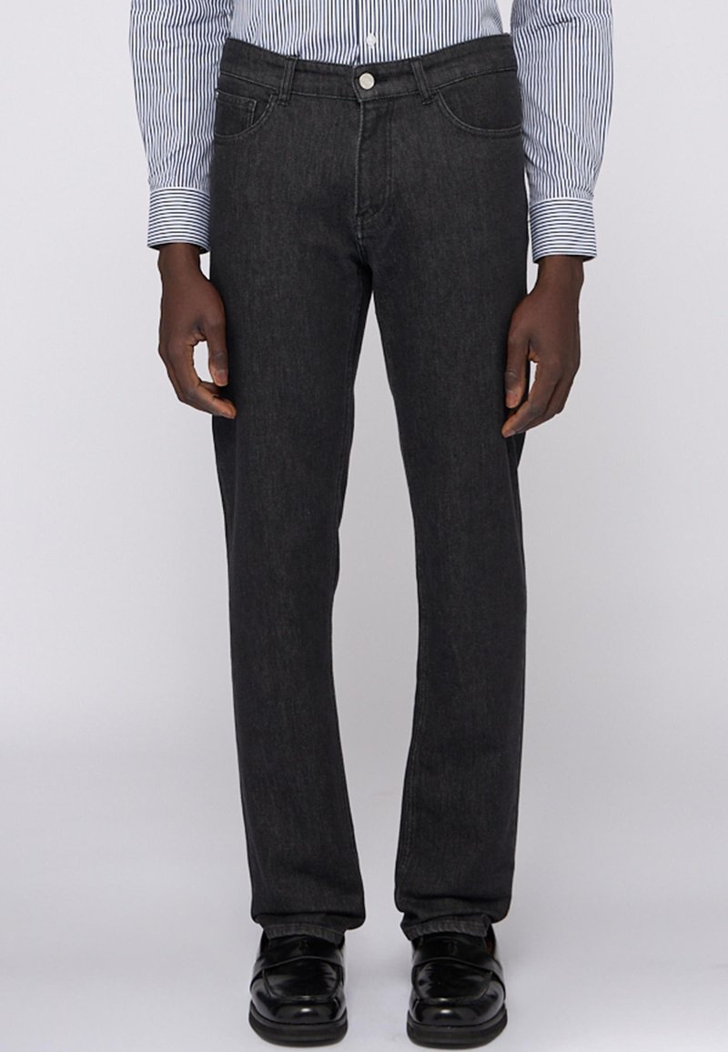 Jeans de mezclilla oscura con un diseño de pierna recta, cinco bolsillos y cierre frontal con botón. Llevados con zapatos de vestir negros brillantes.