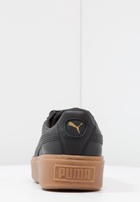 Sneaker nera in pelle con superficie testurizzata, suola piatta color beige e logo dorato Puma sul tallone.