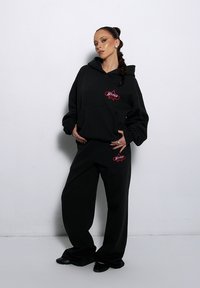 Czarna bluza z kapturem i pasujące dresy ze czerwonymi haftowanymi akcentami. Luźny krój i miękka tekstura, z kieszenią typu kangur.