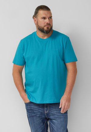 Baarddragende man met naar achteren gekamd haar, gekleed in een felblauw T-shirt met korte mouwen en een blauwe spijkerbroek, staand tegen een effen grijze achtergrond.