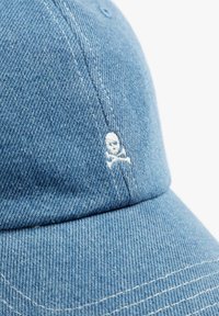 Denim-blaues Cap mit gesticktem weißen Totenkopf- und Knochenmotiv. Abgerundete, weiche Krone mit sichtbaren Nahtlinien und gesticktem Schirm.