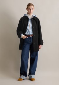 Zwarte wollen jas, blauwgestreept shirt, hoogstaande blauwe spijkerbroek met opgerolde mouwen, bruine leren riem en beige schoenen. Neutrale achtergrond.