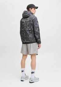 Mand iført sort kasket, sort mønstret hættetrøje med teksten "CHASE THE FIRST LIGHT", lysegrå shorts, hvide sokker og grå sneakers, som står.