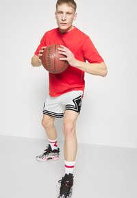 Rotes T-Shirt mit kurzen Ärmeln, weiße Basketballshorts mit schwarzen Akzenten, hält einen Spalding-Basketball, trägt gemusterte schwarze Sneaker und weiße Socken.