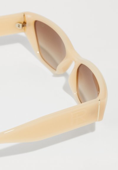 Beige sunglasses with a glossy finish, gradient brown lenses, angular design, and a logo embossed on the frame's side.