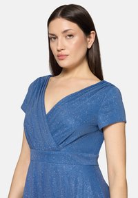 Femme aux cheveux longs et raides, vêtue d'une robe portefeuille bleu scintillante à manches courtes, sur un fond clair uni.