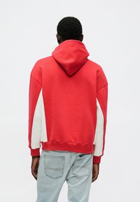 Felpa rossa con pannello laterale bianco, dotata di tasca anteriore a marsupio e cappuccio regolabile con coulisse. Tessuto morbido in misto cotone.