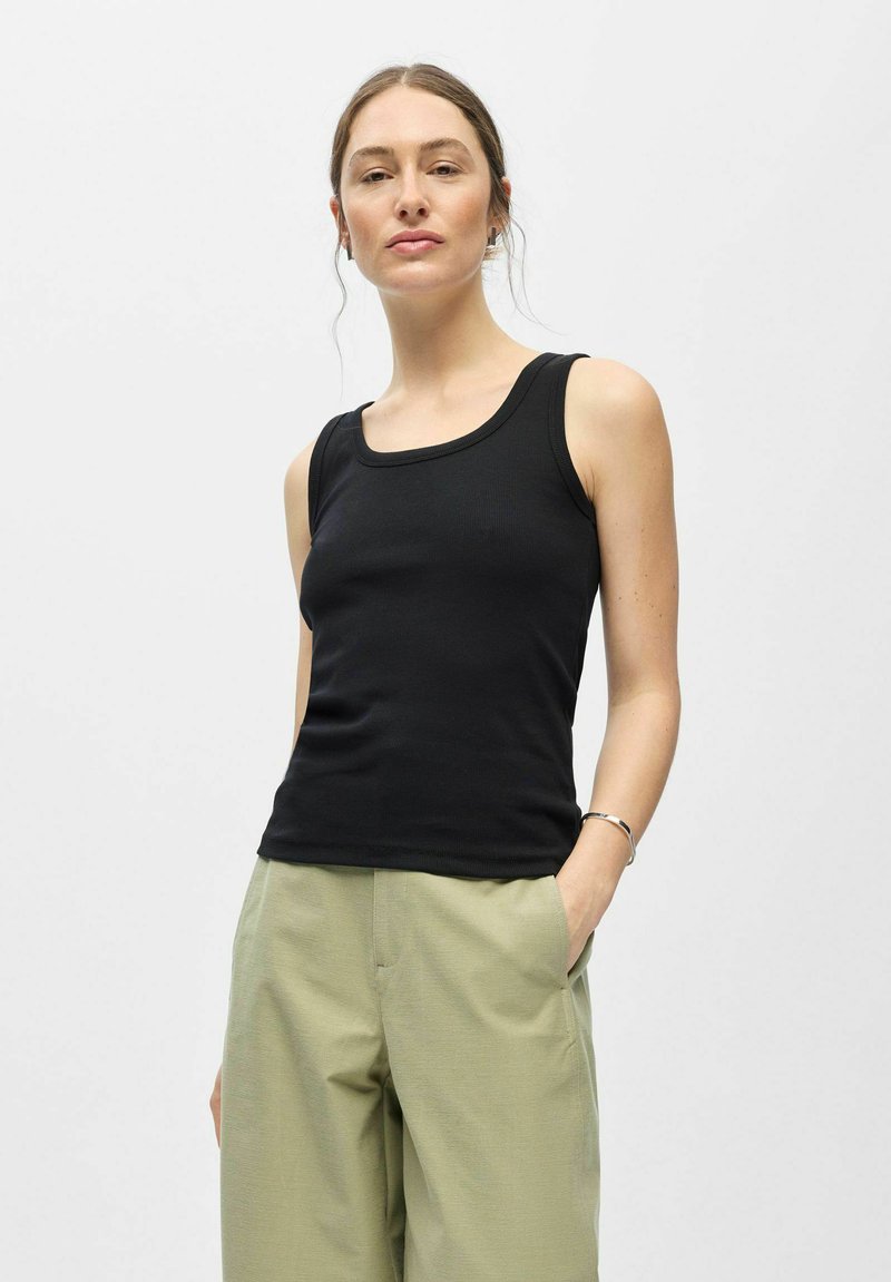 Jeune femme à la peau claire et aux cheveux bruns attachés, portant un haut noir sans manches et un pantalon vert clair, debout avec une main dans la poche.