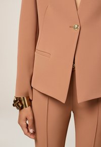 Blazer su misura rosa chiaro con bottone dorato, abbinato a pantaloni coordinati. Presenta una tasca singola e un tessuto liscio e texture.