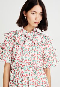 Blusa blanca con estampado de cerezas rojas, mangas con volantes y un lazo atado en el cuello. Tejido ligero con una silueta femenina y cierre de botones.