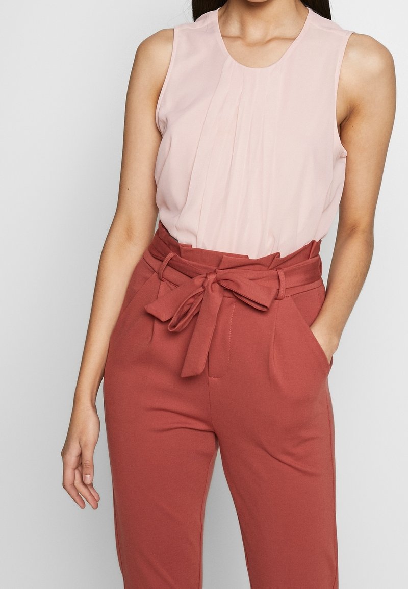 Femme portant un haut sans manches rose clair rentré dans un pantalon taille haute couleur rouille avec une ceinture en tissu nouée et les mains dans les poches.