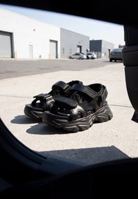 Paire de sandales à plateforme épaisses noires avec des brides en velcro posées sur un trottoir près de voitures garées et de bâtiments industriels.