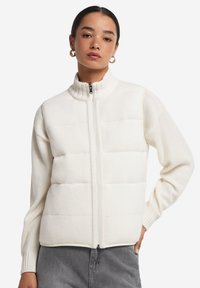 Weiße Zip-Jacke mit hohem Kragen, ausgestattet mit einem strukturierten Strickoberteil und einem gepolsterten Unterteil. Glatte Oberfläche und gerippte Bündchen.