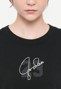 Melns T-krekls ar apaļu dekoltē, ar baltu izšūtu "Jordan" parakstu un numuru "23" pelēkā ēnu dizainā.